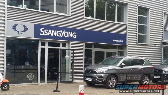 Gray SsangYong Rexton SUV parked in front of a dealership, showing "Rexton" text on the door