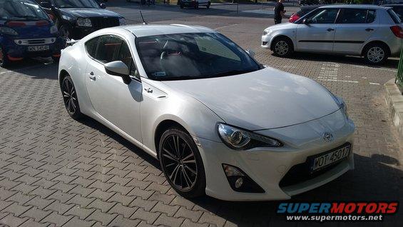 White sports coupe parked on brick pavement. Other cars visible in background