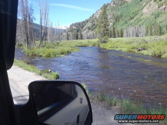 Side mirror reflection of a white vehicle showing a mountain stream scene