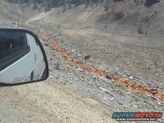 White van side mirror showing reflection of a dry, rocky landscape with an orange-stained streambed