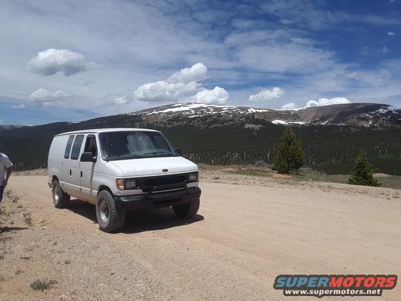 1999 Ford E250 van parked on a dirt road, mountains and cloudy sky behind it