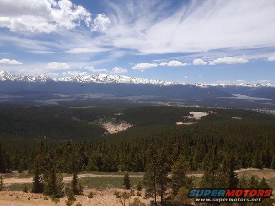 Mountain range with snow-capped peaks overlooks a vast forest and valley under a blue sky with clouds. A dirt road is visible in the foreground