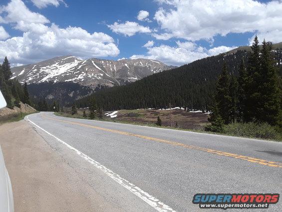 Winding asphalt road with double yellow lines curves through a mountain landscape with snow-capped peaks and pine forests under a cloudy blue sky