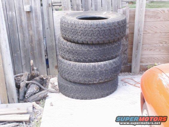 stack of four off-road tires leaning against a wooden fence