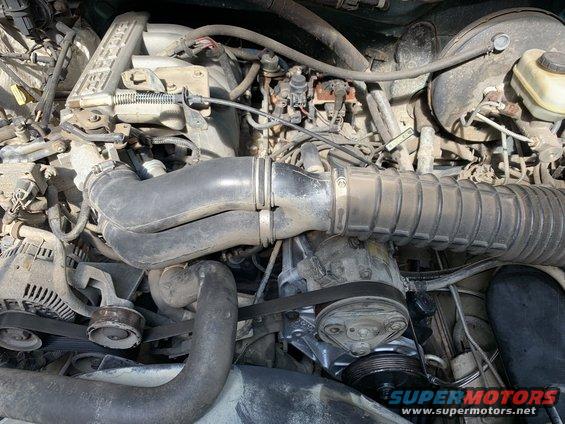 Engine bay with hoses, belts, and engine components visible. Some dust and grime present