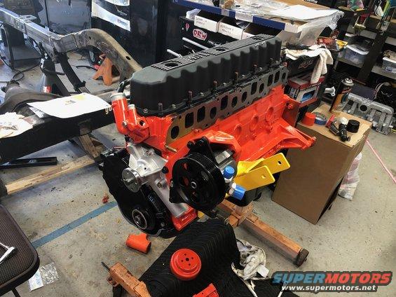 Orange engine block with black valve cover on a wooden stand, with a partial vehicle frame in the background