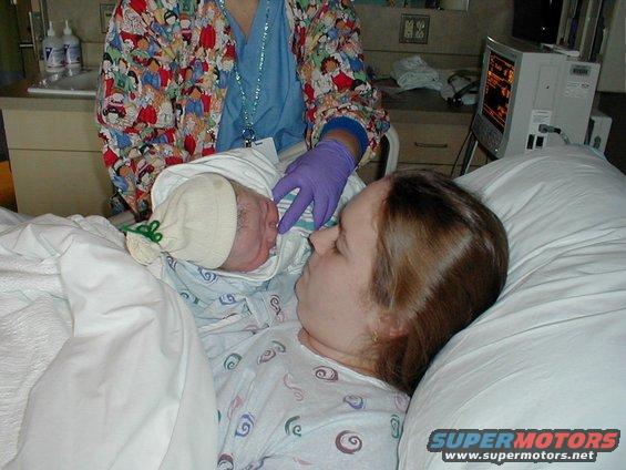 Newborn baby wrapped in a blanket and hat, held by a nurse, with mother looking on