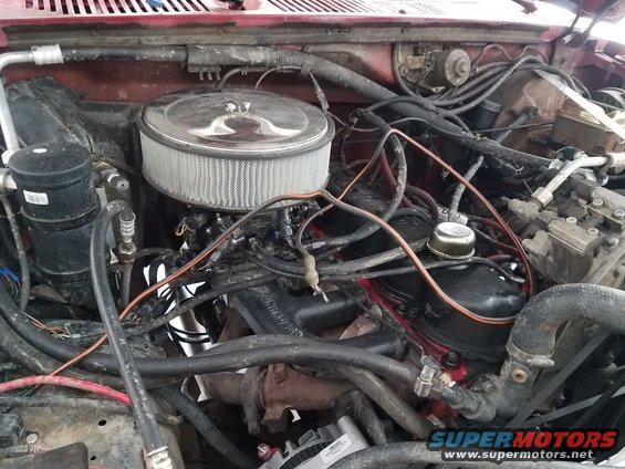 Engine bay of a red vehicle, showing a V8 engine with an aftermarket air filter and various hoses and wires. The condition appears used
