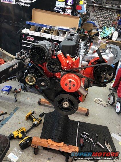 Engine assembly on a stand with tools and parts scattered around. Includes red painted components and black belts