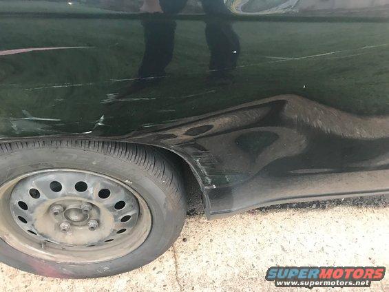 Dark green car's rear quarter panel with significant dents and scratches, next to a steel wheel with a flat tire