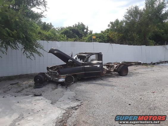 Wrecked black truck chassis with hood open, missing body panels and wheels. Appears to be from the 1950s