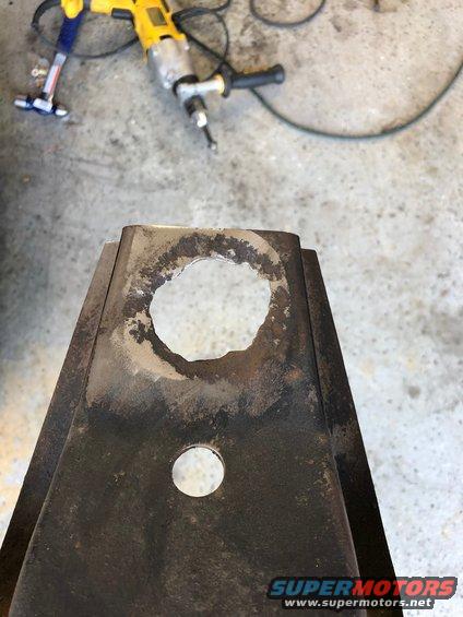 Close-up of a rusty metal part with a large hole drilled through it. A yellow drill is visible in the background
