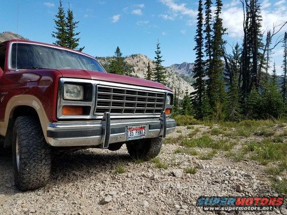 Red 1985 Ford Bronco front view parked on a gravel road with mountains in background