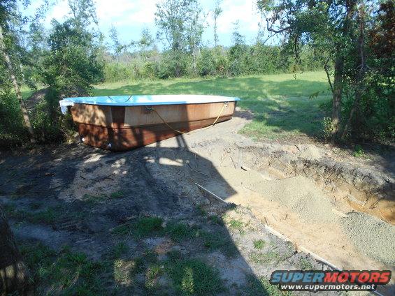 Above-ground pool with blue liner and brown exterior walls sits on prepared ground next to a dug pit