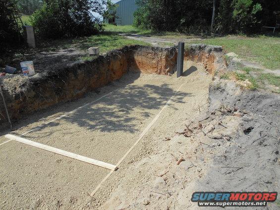 excavated hole with gravel base and wooden markers, pipe sticking out of ground