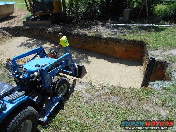 Blue tractor with front loader, yellow excavator, and worker operating a compactor at a construction site