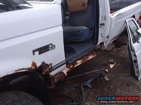 White pickup truck with extensive rust on the lower body and door, showing the interior driver's seat and door panel