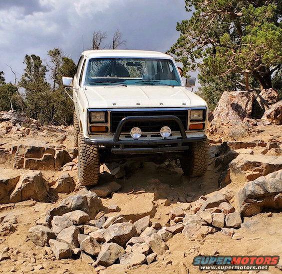 White Ford Bronco navigating a rocky trail, front view. The grille and body style suggest an early 1980s model