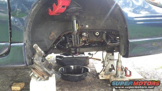 Car's front suspension disassembled with red brake calipers, engine components visible, and oil drain pans on the ground