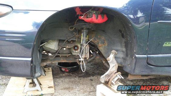 Dark blue car's front wheel assembly removed, with red brake caliper and oil pan visible