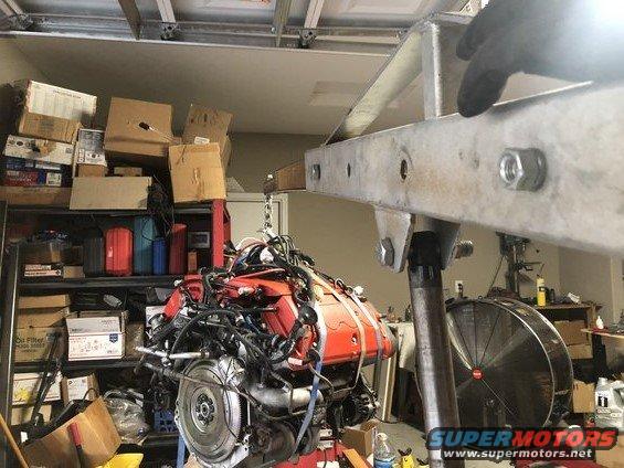 Engine assembly on a red stand, suspended by a metal hoist in a cluttered garage
