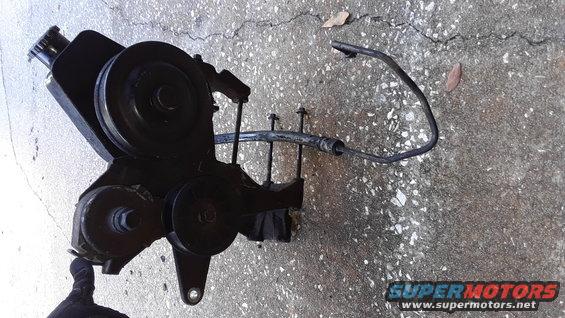 Power steering pump assembly with pulleys and hoses, lying on concrete