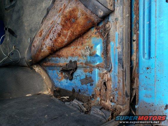 Interior door frame and floor of a vehicle showing significant rust damage and torn upholstery