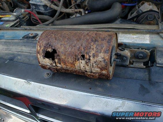 Heavily rusted cylindrical exhaust part resting on a car's hood, with engine components visible in the background