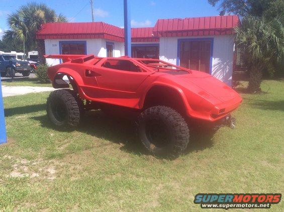 Red custom vehicle with large off-road tires and a body resembling a Lamborghini Countach, parked on grass