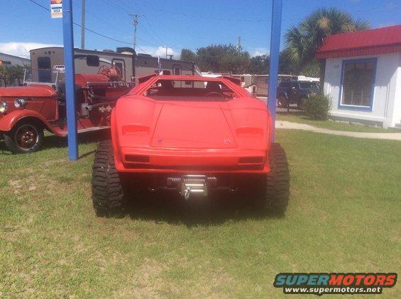 Red kit car with large tires and a winch on the front, parked on grass next to a vintage fire truck