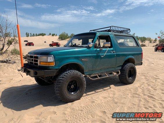 1996 Ford Bronco in teal with roof rack and off-road tires parked on sand dunes, other vehicles in background