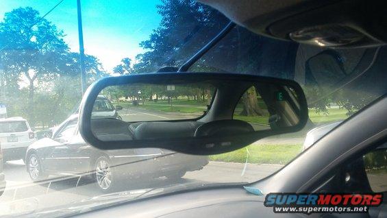 Interior rearview mirror shows traffic on a road, including a dark sedan and a white SUV