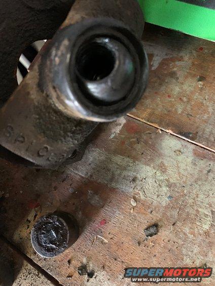 Close-up of a rusty brake caliper part with a threaded hole. A small cap with grease sits on a wooden surface