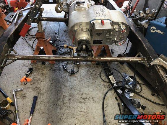 Vehicle frame on jack stands with a large metal component, possibly a transmission or transfer case, suspended above
