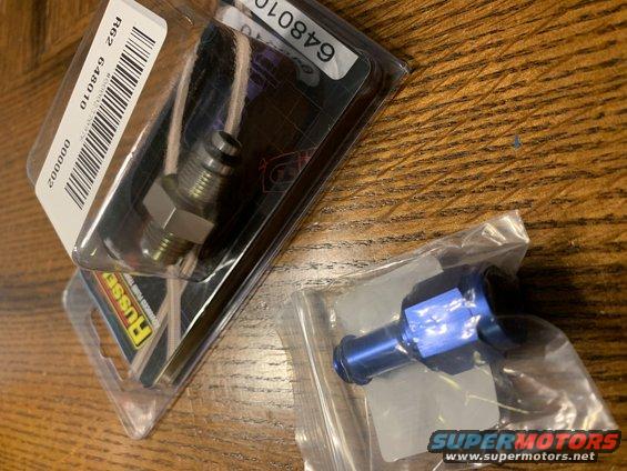 Two automotive fittings in packaging on a wooden surface. One is a metal fitting in a clear blister pack with a barcode and part number R62 648010. The other is a blue anodized fitting in a plastic bag with a Supermotors logo