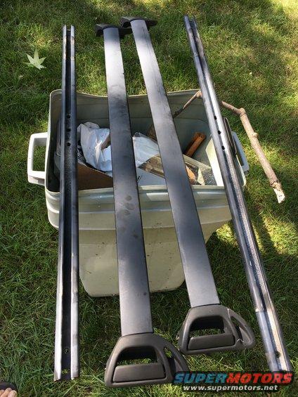 Two roof rack crossbars and two side rails laid out on grass next to a storage bin