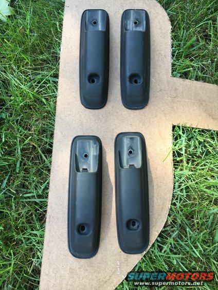 Four black plastic car door handles laid out on cardboard with grass in background. Supermotors.net logo visible