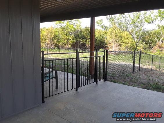 Backyard patio area with a chain-link fence and gate, leading to trees and brush
