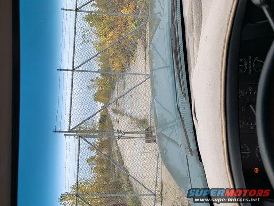 Dashboard view from inside a vehicle looking out through the windshield at a chain-link fence and trees. Part of the dashboard and gauges are visible on the right