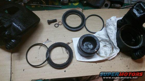 Disassembled brake caliper and seals on a workbench. Includes caliper housing, piston, O-rings, and a bolt