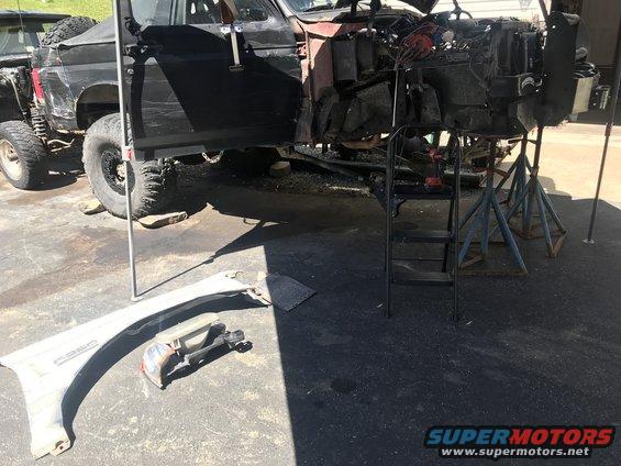 Black Ford Bronco body on jack stands, engine bay open, detached fender and headlight on ground