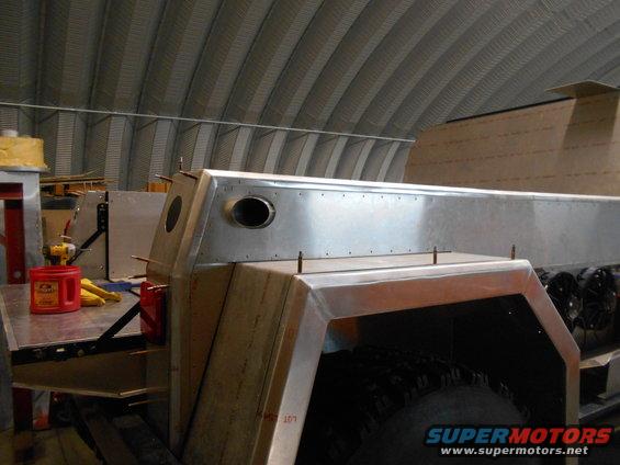 Unfinished metal body of a custom vehicle, showing exposed rivets and a circular vent. A red container and yellow items are on a workbench