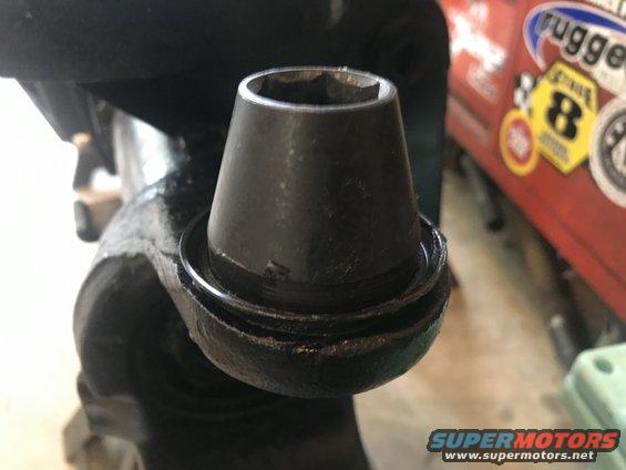 Close-up of a black automotive ball joint with a socket on top. Red vehicle with stickers visible in background
