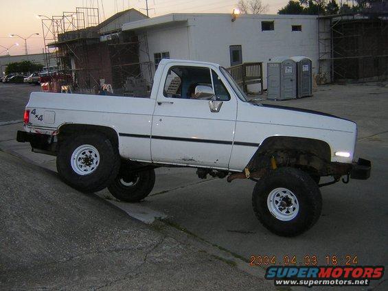 White pickup truck with 4x4 badging, lifted suspension, and off-road tires, angled on a concrete curb