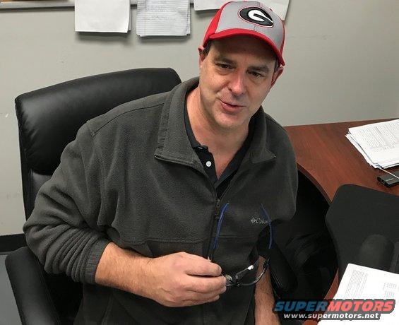 Man wearing a red and gray baseball cap sits in an office chair, holding eyeglasses