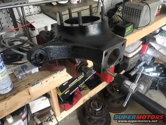 Black painted vehicle axle housing assembly in a vise with a tool inserted into a bolt hole