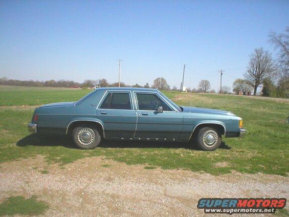 Blue sedan parked on gravel with a grassy field behind it. The car has a two-tone paint job and chrome hubcaps