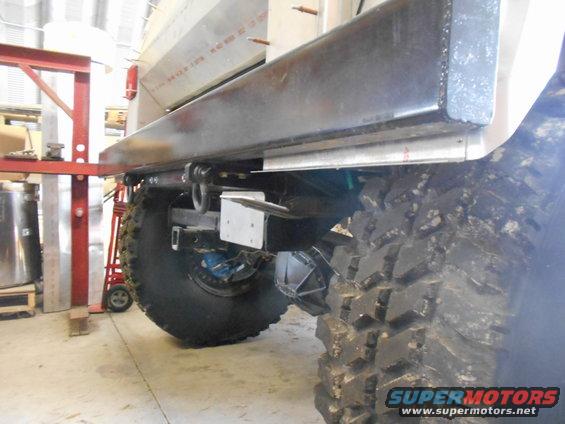 Close-up of a large, knobby tire and the rear axle assembly of a heavily modified off-road vehicle, with a metal bed frame above