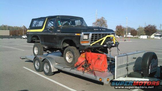 Black 1978 Ford Bronco with yellow trim on a trailer, lifted with off-road tires, carrying a red snowblower