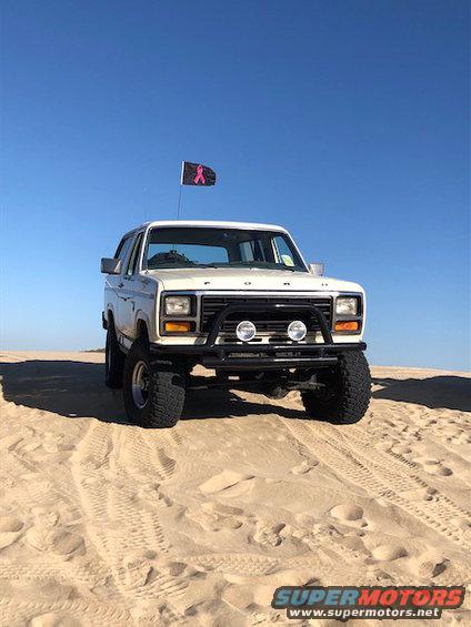 White 1981 Ford Bronco with bull bar and pink ribbon flag on sand dune